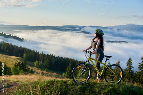 Sporty girl cyclist riding on yellow bicycle on a rural trail in the mountains, wearing helmet, enjoying valley view on sunny morning. Foggy mountains, forests on the blurred background