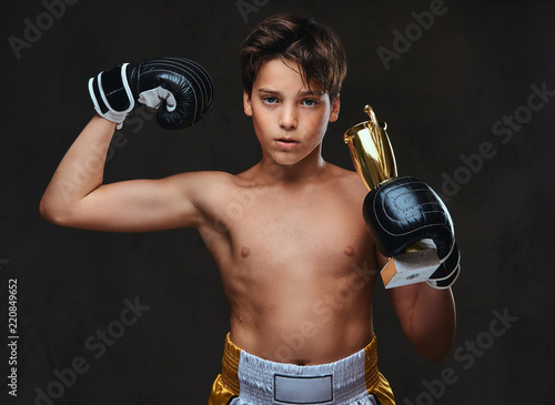 Young shirtless boxer champion wearing gloves holds a winner's cup showing muscles.