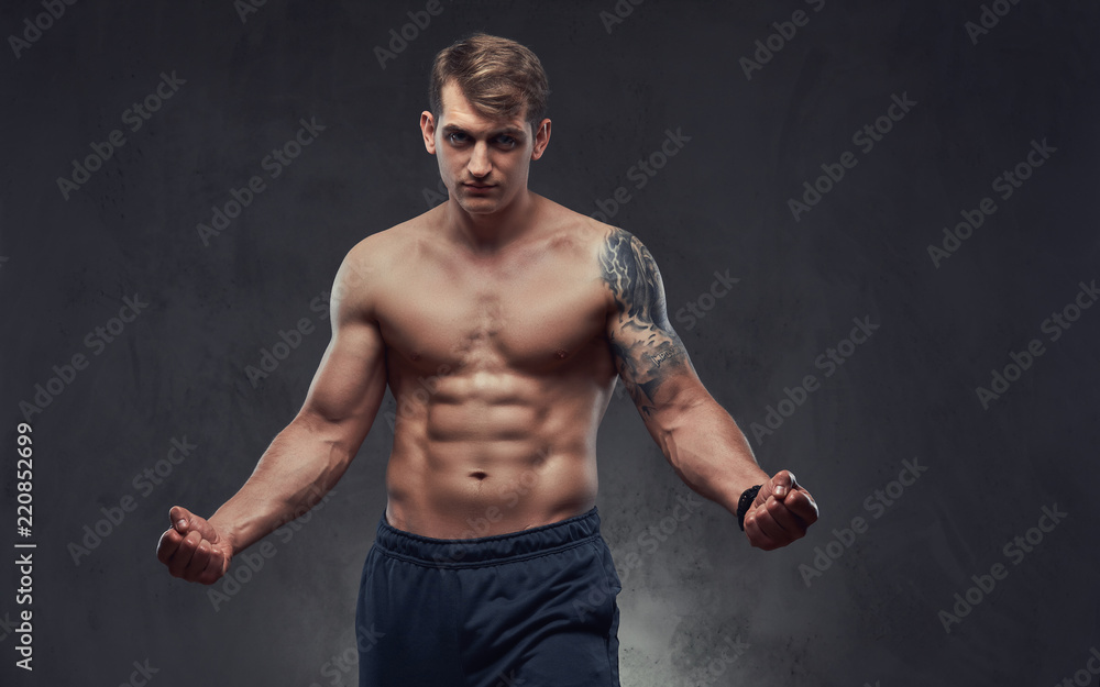 Young handsome shirtless sportsman with muscular torso and tattoo on his hand pose in a studio on a dark textured background.