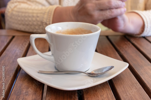 Cup of coffee on a wooden table and hands of an elderly woman behind.