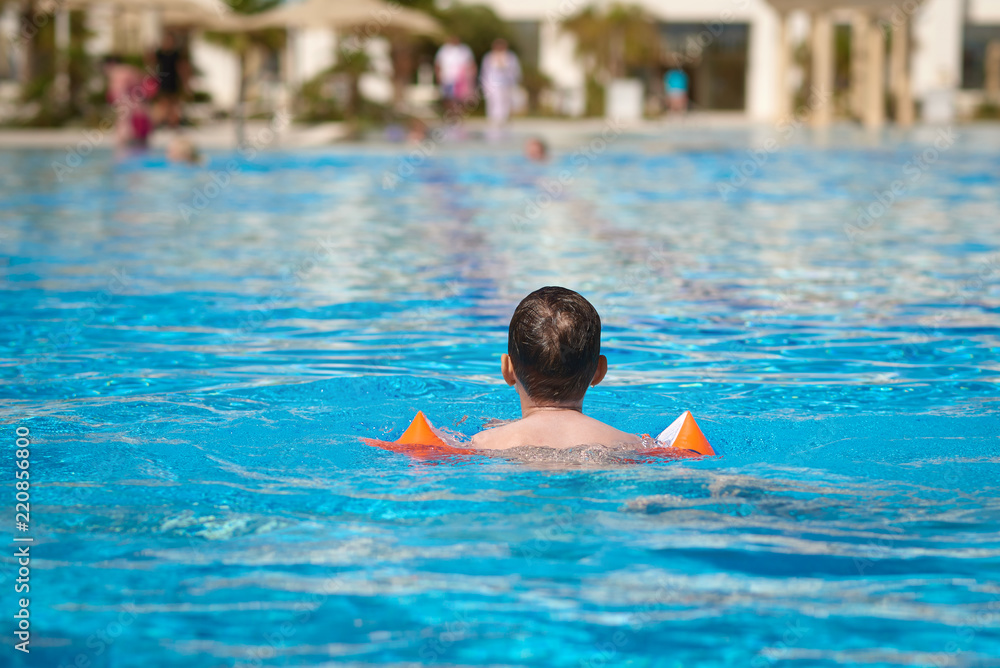European boy in floating sleeves swimming in pool in summer. Back view.