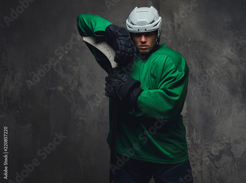 Hockey player wearing green protective gear and white helmet standing with the hockey stick on a gray background. photo