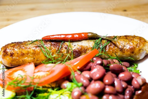 On a white plate with a serving option lies a delicious meal. Spices are sprinkled with fresh cucumbers, red beans, tomatoes, dill and fried in a skillet, a natural sausage, which is decorated with ch photo