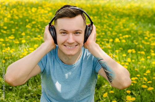 Portrait of a young guy listening to music on headphones and sitting on green grass with dandelions.