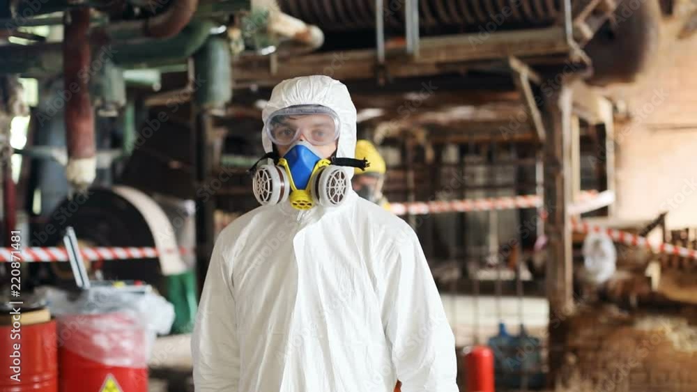 Caucasian scientist in new white protective suit looking up. Portrait of serious Slavic young man in respirator mask. Abandoned nuclear power station. Caucasian scientist in new white protective suit looking up. Portrait of serious Slavic young man in respirator mask. Abandoned nuclear power station.