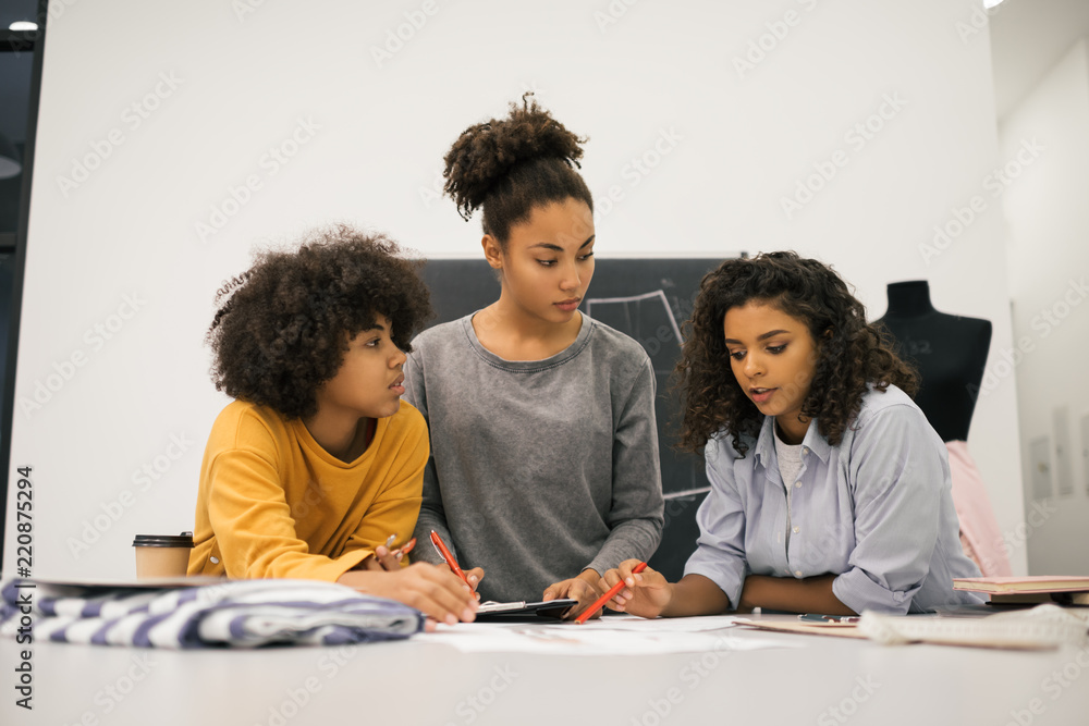 Creative team of stylish African American women planning new startup project in workplace. Brainstorming. University students learning fashion design at atelier. Young hipsters working together.