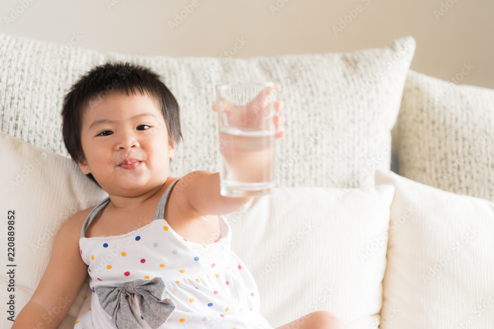 Happy little asian girl hand holding drinking water glass and laughing, sitting on sofa at home. Medicine and health care concept. Happy little asian girl hand holding drinking water glass and laughing, sitting on sofa at home. Medicine and health care concept.