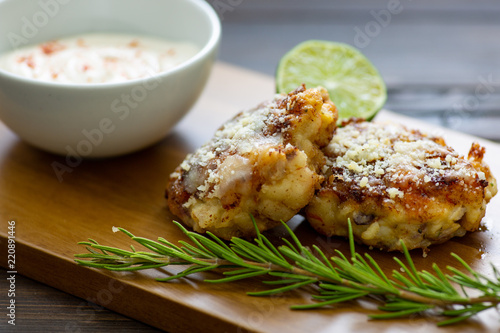 Two shrimp cakes on a cutting board with a lemon garlic dipping sauces lemon wedge and a stem or rosemary on a wooden table.