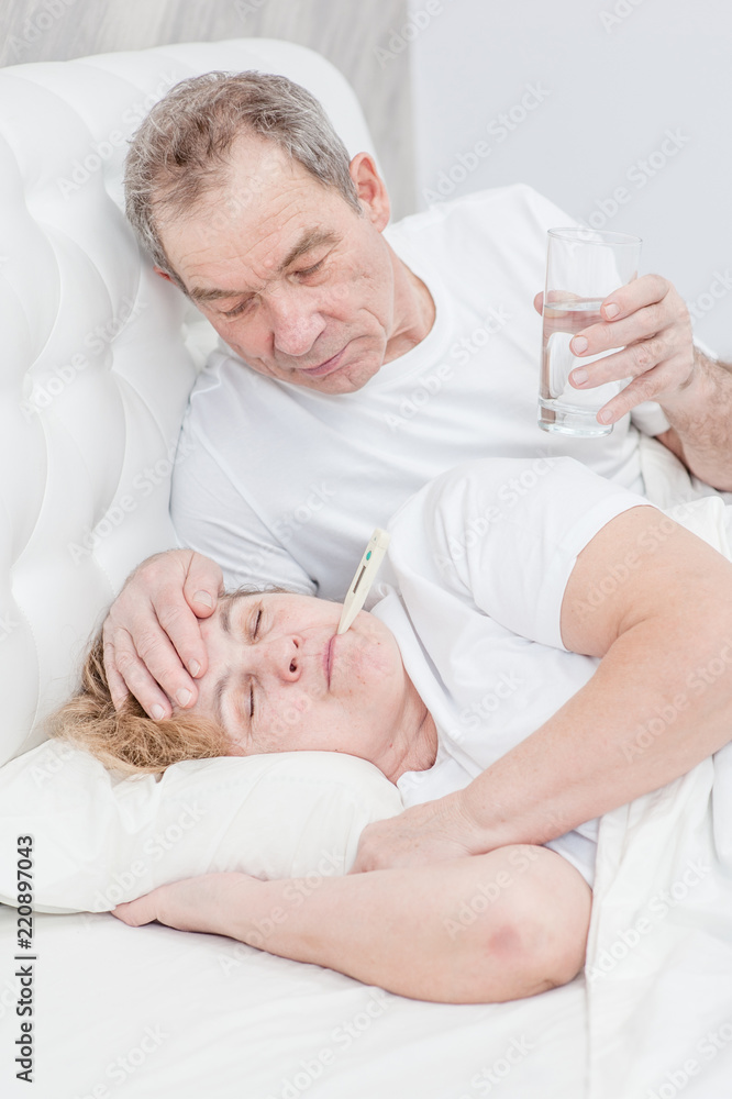 an elderly man with glass of the water touches forehead his wife, checking the temperature an elderly man with glass of the water touches forehead his wife, checking the temperature