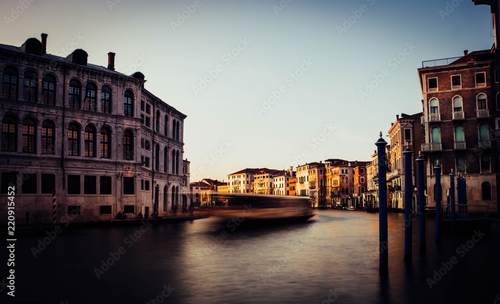 A boat in motion crosses the Grand Canal of Venice at sunset, while the sun hits the face of some historic buildings on the banks of the canal