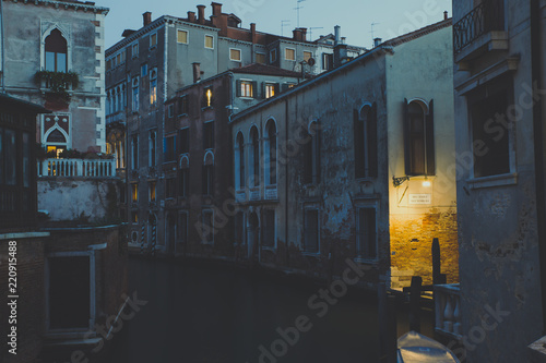 View of a Venice canal at dusk, while the faint light coming from the street lamps starts to illuminate the water creating a sort of magical effect in this city