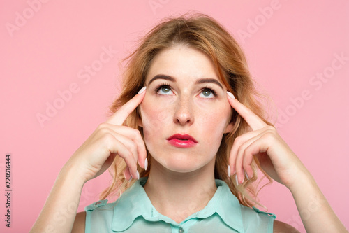 mind games telepathy thought transfer and brain power. beautiful girl concentrating on invisible object above her. index fingers on temples. young beautiful blond woman portrait on pink background.