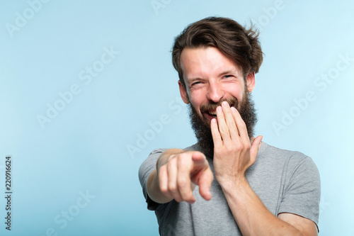 omg. ha-ha. bearded hipster man mocking and laughing at viewer pointing finger and covering mouth. sneer humiliation and psycological abuse concept. portrait of a grinning guy on blue background. photo
