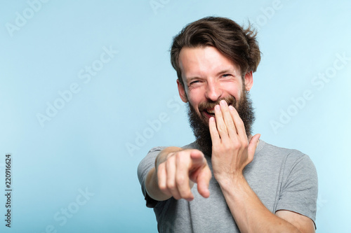 omg. ha-ha. bearded hipster man mocking and laughing at viewer pointing finger and covering mouth. sneer humiliation and psycological abuse concept. portrait of a grinning guy on blue background.