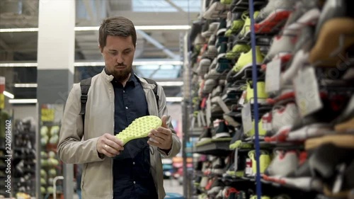Man shopper is watching pair of shoes in sport store. He is checking quality and taking second sneaker from shelf, thinking about buying