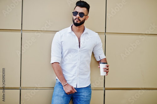 Stylish tall arabian man model in white shirt, jeans and sunglasses posed at street of city. Beard attractive arab guy with cup of coffee against golden wall.