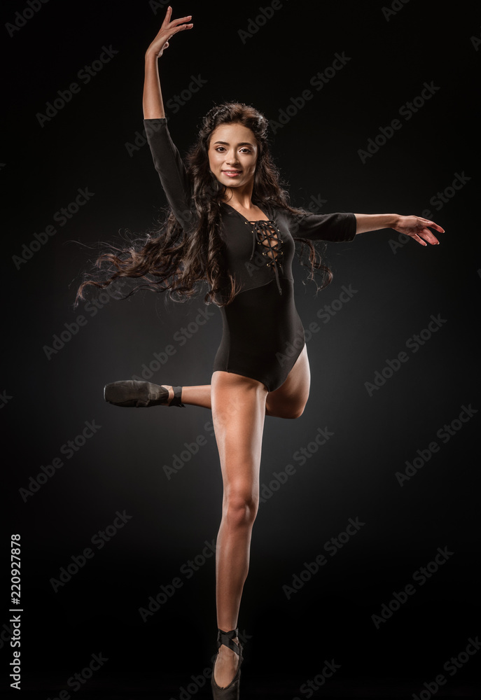 smiling ballerina in black bodysuit and ballet shoes dancing on dark backdrop