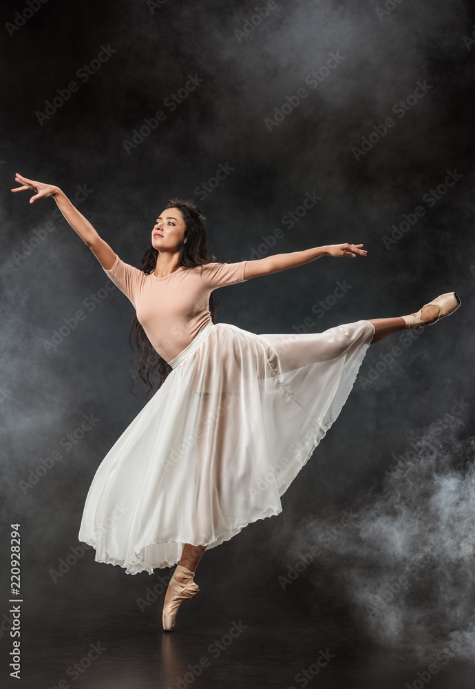 beautiful young ballerina in white skirt dancing on dark background with smoke around