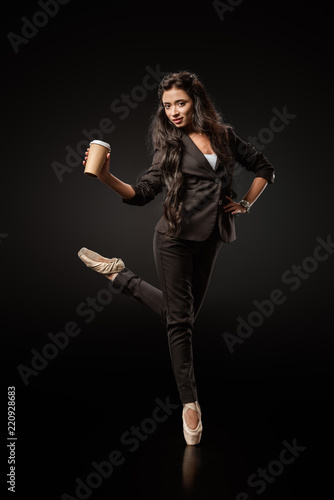 attractive young businesswoman in formal wear and ballet shoes with coffee to go on black background