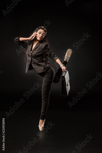 stylish businesswoman in formal wear and ballet shoes talking on smartphone on black backdrop