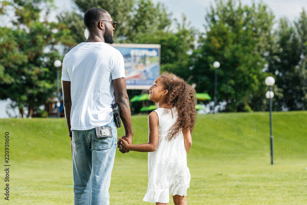 rear view of african american police officer and daughter holding hands and walking in amusement park rear view of african american police officer and daughter holding hands and walking in amusement park
