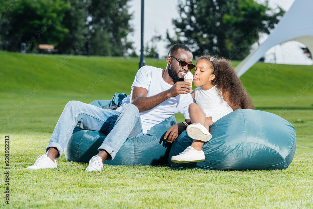 african american father giving ice cream to daughter on beanbag chairs in park african american father giving ice cream to daughter on beanbag chairs in park