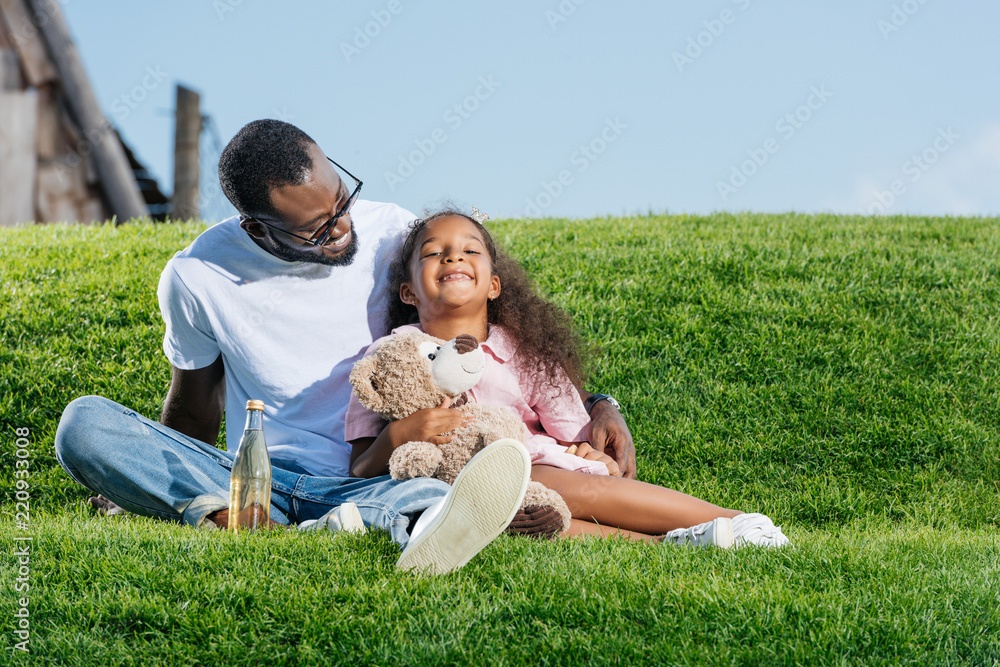 african american father and smiling daughter sitting on hill with soda and teddy bear at amusement park african american father and smiling daughter sitting on hill with soda and teddy bear at amusement park