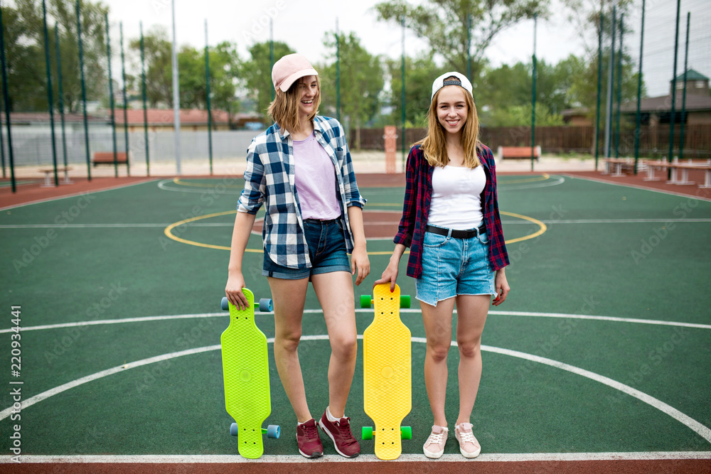 Obraz premium Two pretty smiling blond girls wearing checkered shirts, caps and denim shorts are standing on the sportsfield with bright longboards in their hands. Beautiful legs in a good fit. 