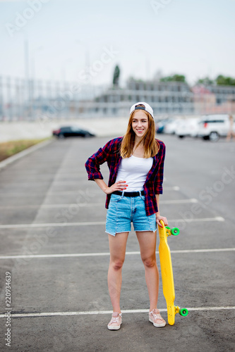 A beautiful blond girl wearing checkered shirt, cap and denim shorts is standing on the car park with a yellow longboard in her hand. Sport and cool style.