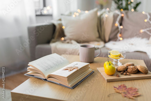 hygge and cozy home concept - book, autumn leaves, cup of tea with lemon, almond nuts and oatmeal cookies on table photo