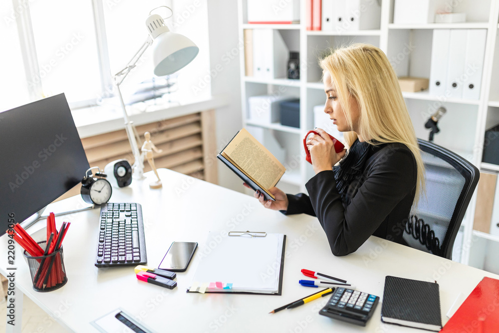 A young girl is sitting at a table in the office, holding a cup and reading a book. A young girl is sitting at a table in the office, holding a cup and reading a book.