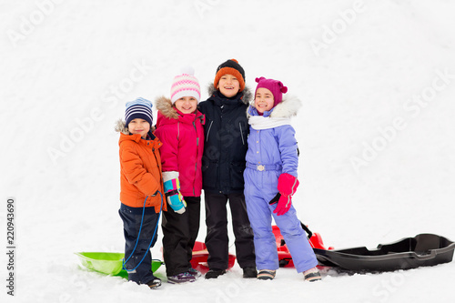 childhood, friendship and season concept - group of happy little kids with sleds hugging in winter