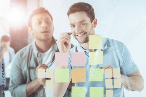 smiling young businessman writing on sticky notes while shocked colleague standing behind © LIGHTFIELD STUDIOS