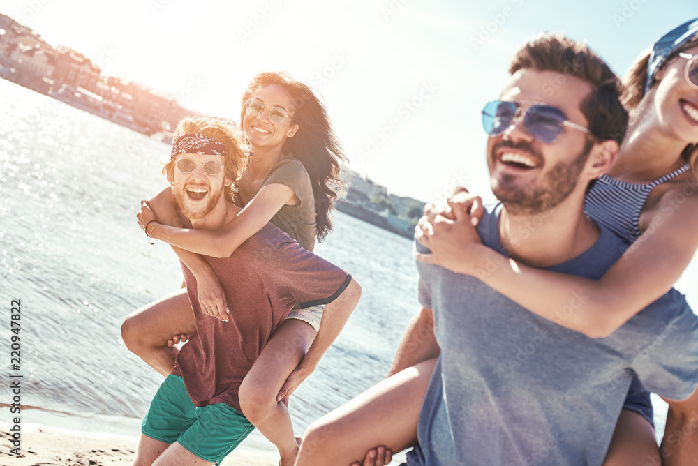Outdoors photo of happy boyfriends piggybacking their girlfriends at sunset on beach