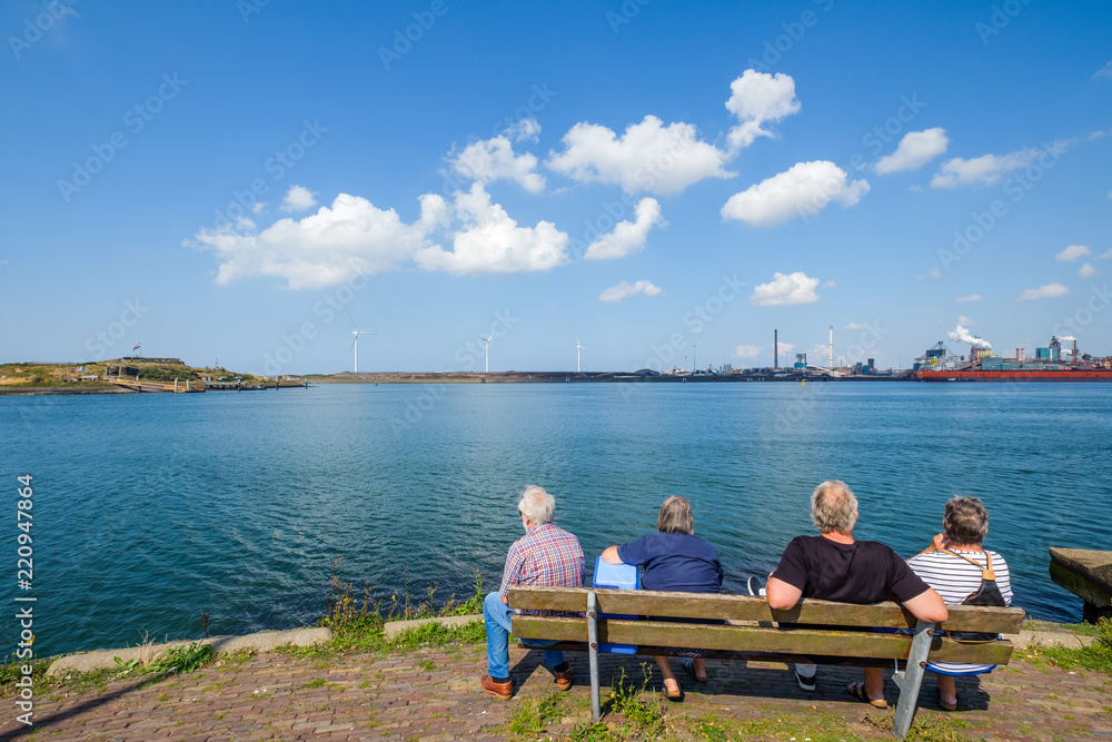Retired people look across the water on the North Sea coast to modern wind turbines and steel works.