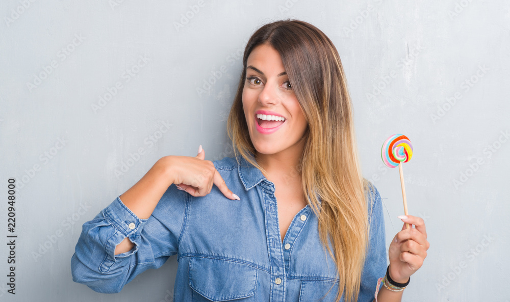 Young adult woman over grey grunge wall eating lollipop candy with surprise face pointing finger to himself Young adult woman over grey grunge wall eating lollipop candy with surprise face pointing finger to himself