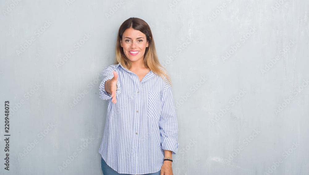 Young adult woman over grey grunge wall wearing fashion business outfit smiling friendly offering handshake as greeting and welcoming. Successful business. Young adult woman over grey grunge wall wearing fashion business outfit smiling friendly offering handshake as greeting and welcoming. Successful business.