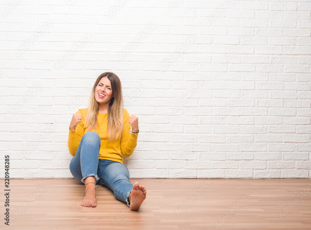 Young adult woman sitting on the floor over white brick wall very happy and excited doing winner gesture with arms raised, smiling and screaming for success. Celebration concept. Young adult woman sitting on the floor over white brick wall very happy and excited doing winner gesture with arms raised, smiling and screaming for success. Celebration concept.