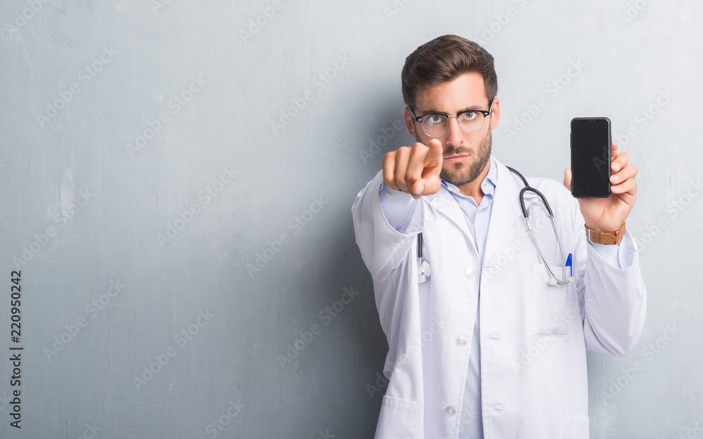 Handsome young doctor man over grey grunge wall holding smartphone pointing with finger to the camera and to you, hand sign, positive and confident gesture from the front Handsome young doctor man over grey grunge wall holding smartphone pointing with finger to the camera and to you, hand sign, positive and confident gesture from the front