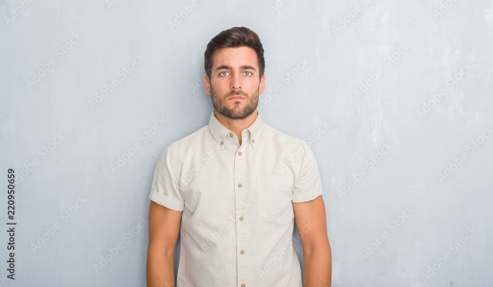 Handsome young man over grey grunge wall wearing summer shirt with serious expression on face. Simple and natural looking at the camera.