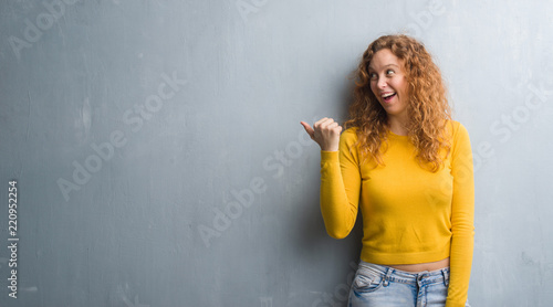 Young redhead woman over grey grunge wall smiling with happy face looking and pointing to the side with thumb up.