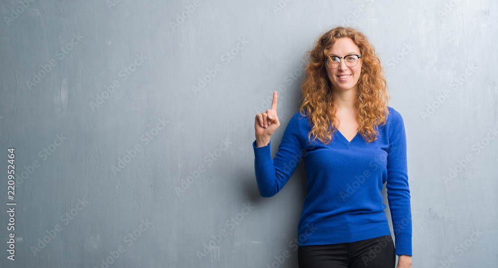 Young redhead woman over grey grunge wall showing and pointing up with finger number one while smiling confident and happy. Young redhead woman over grey grunge wall showing and pointing up with finger number one while smiling confident and happy.