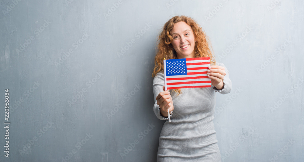 Young redhead woman over grey grunge wall holding flag of United States of America with a happy face standing and smiling with a confident smile showing teeth Young redhead woman over grey grunge wall holding flag of United States of America with a happy face standing and smiling with a confident smile showing teeth