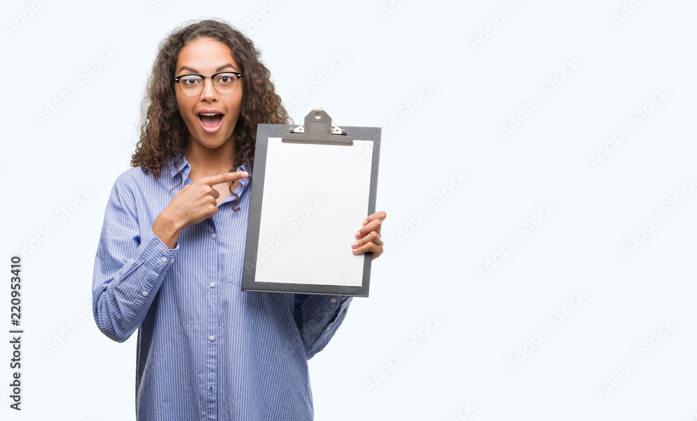 Young hispanic business woman holding clipboard very happy pointing with hand and finger Young hispanic business woman holding clipboard very happy pointing with hand and finger