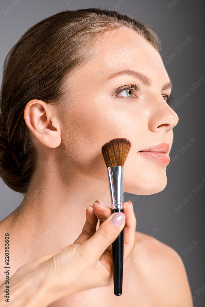 partial view of makeup artist applying blush on models face isolated on grey partial view of makeup artist applying blush on models face isolated on grey