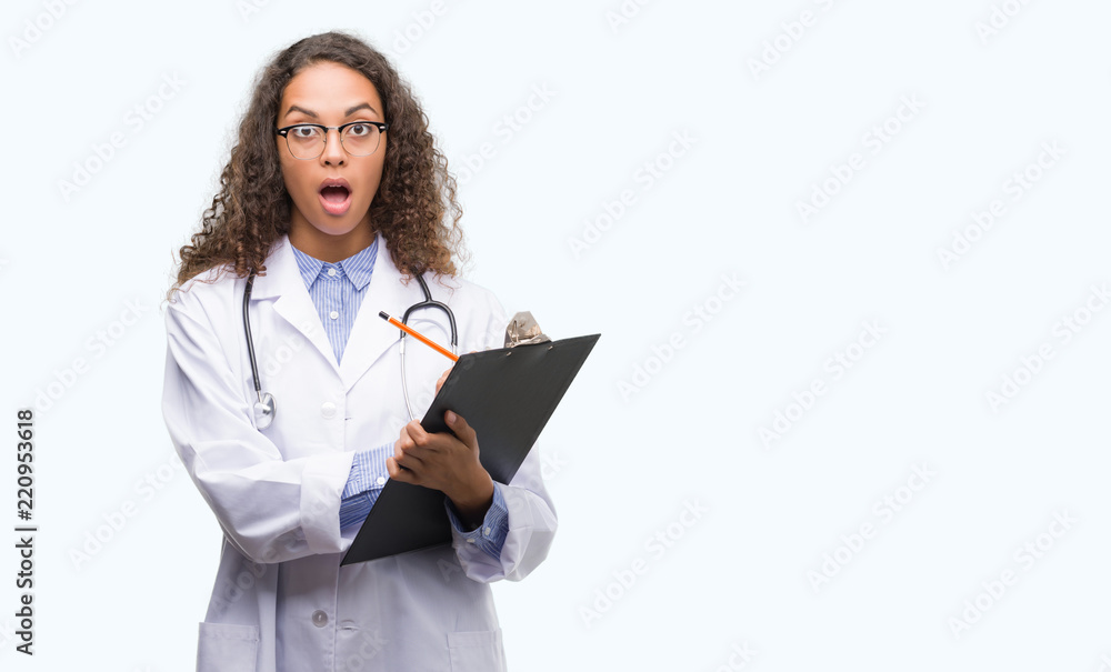 Young hispanic doctor woman holding a clipboard scared in shock with a surprise face, afraid and excited with fear expression Young hispanic doctor woman holding a clipboard scared in shock with a surprise face, afraid and excited with fear expression