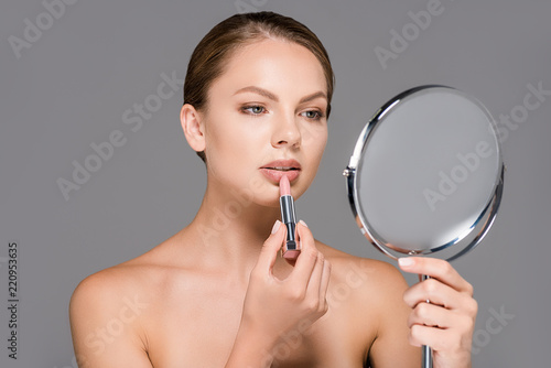 portrait of young woman looking in mirror while applying nude lipstick isolated on grey