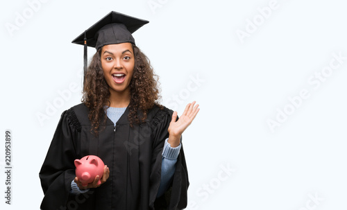 Young hispanic woman wearing graduation uniform holding piggy bank very happy and excited, winner expression celebrating victory screaming with big smile and raised hands