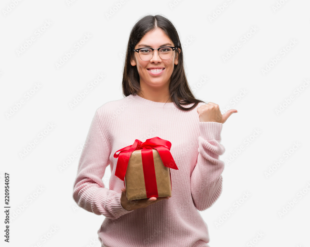 Young hispanic woman holding a gift pointing and showing with thumb up to the side with happy face smiling Young hispanic woman holding a gift pointing and showing with thumb up to the side with happy face smiling
