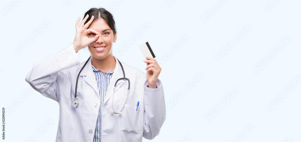 Young hispanic doctor woman holding credit card with happy face smiling doing ok sign with hand on eye looking through fingers Young hispanic doctor woman holding credit card with happy face smiling doing ok sign with hand on eye looking through fingers